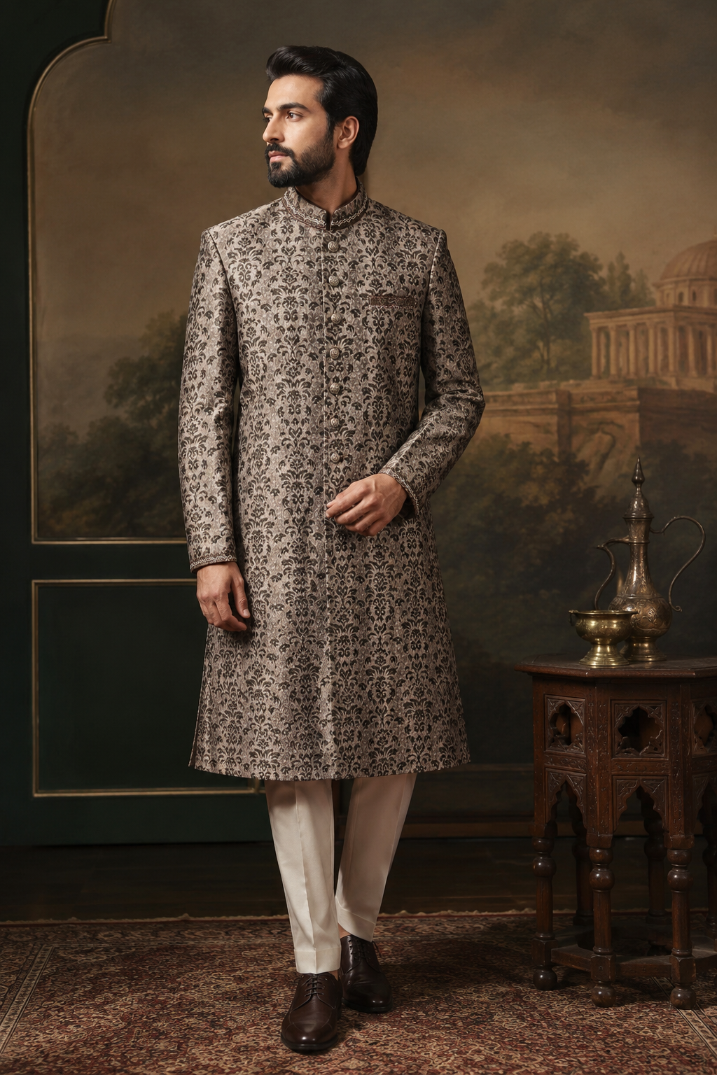 A man wearing a traditional Indian sherwani long coat in a brown and beige patterned fabric, paired with cream trousers and dark brown shoes, standing in a styled room with a painted backdrop and a wooden table.