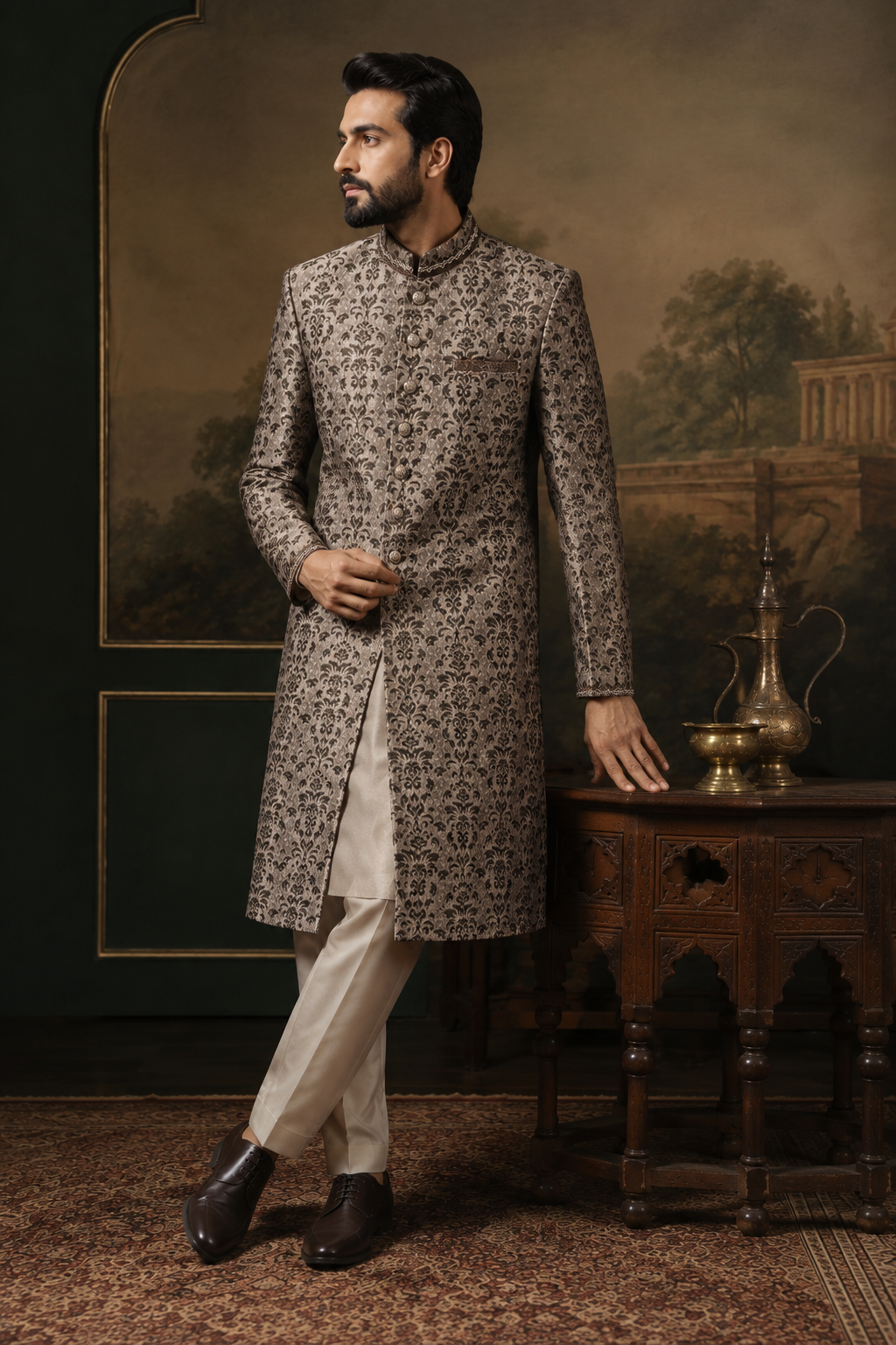 A man wearing a traditional Indian sherwani long coat in a brown and beige patterned fabric, paired with cream trousers and dark brown shoes, standing in a styled room with a painted backdrop and a wooden table.